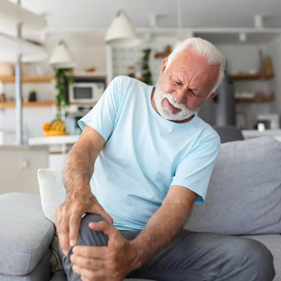 An older man is sitting on the couch at home, and is holding his leg. It looks like he has pain in the knee joint.